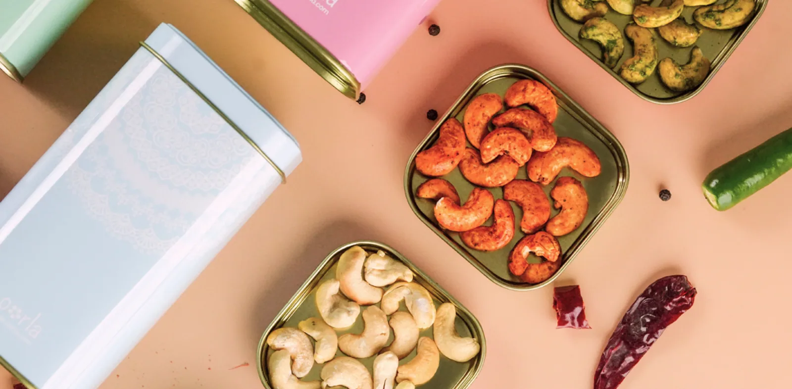 Three open tins of baked cashew — plain, chilli and herb — beside pastel Oorla tins on a peach ground, photographed from above