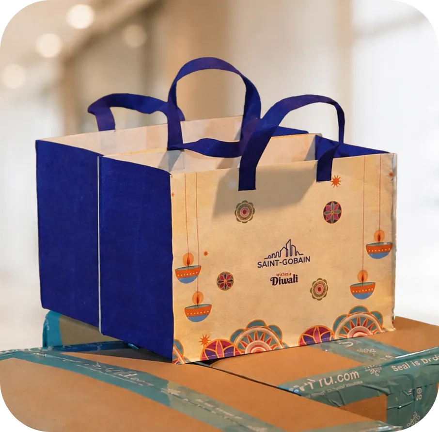A Saint-Gobain Diwali carry bag in cream and blue with rangoli and diya artwork, standing on dispatch cartons